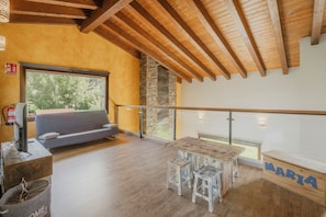 A spacious room with a wooden ceiling, a stone fireplace, a balcony with a glass railing, and a table with stools.