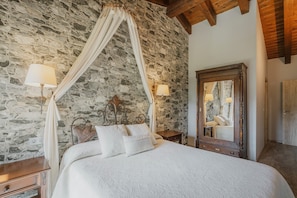 A bedroom with a stone wall, a canopy bed, a wooden headboard, bedside lamps, a mirror, and a wooden nightstand.