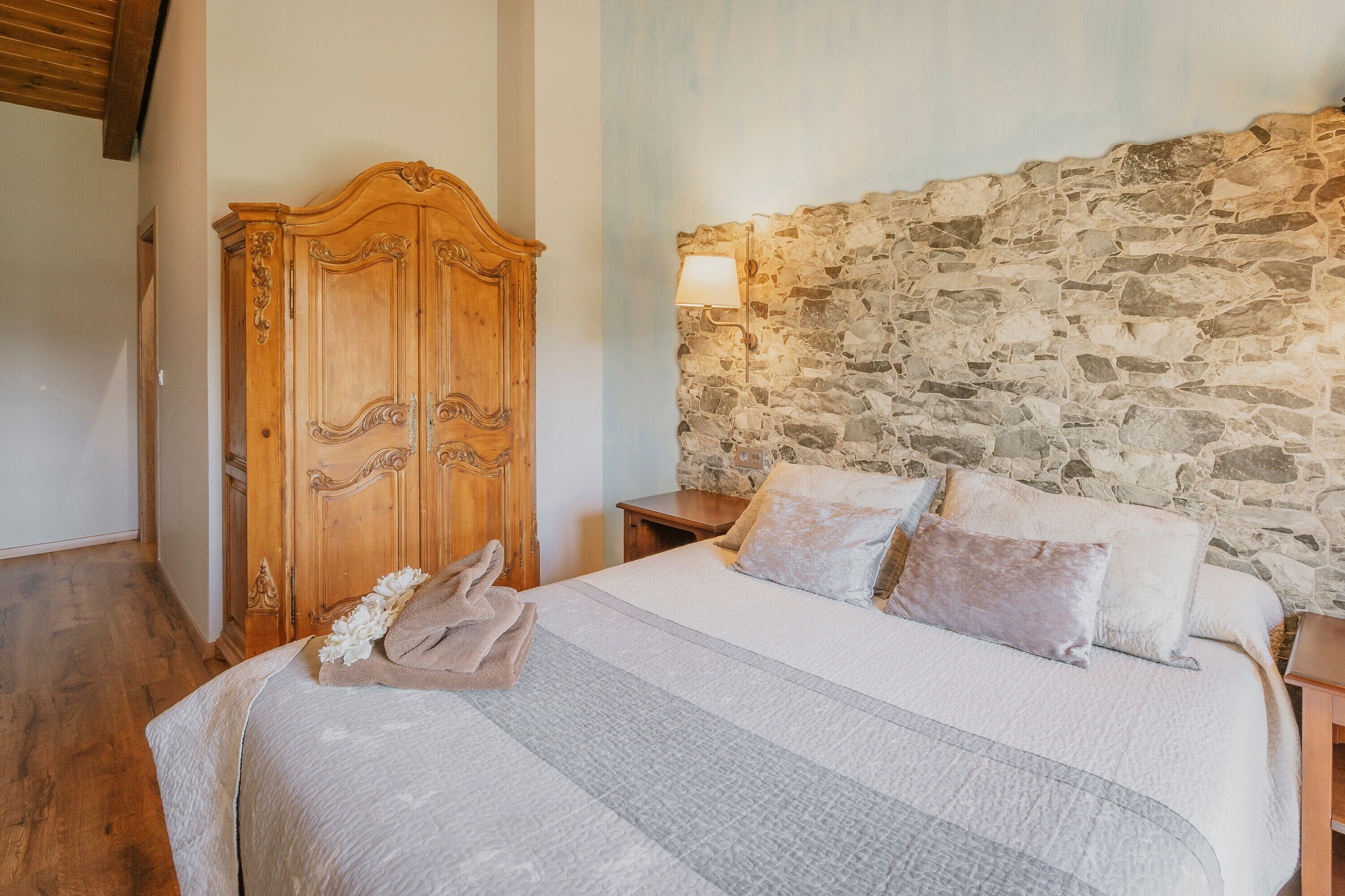 A bedroom with a stone wall, a wooden wardrobe, a bed with pillows, and a nightstand.