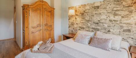 A bedroom with a stone wall, a wooden wardrobe, a bed with pillows, and a nightstand.