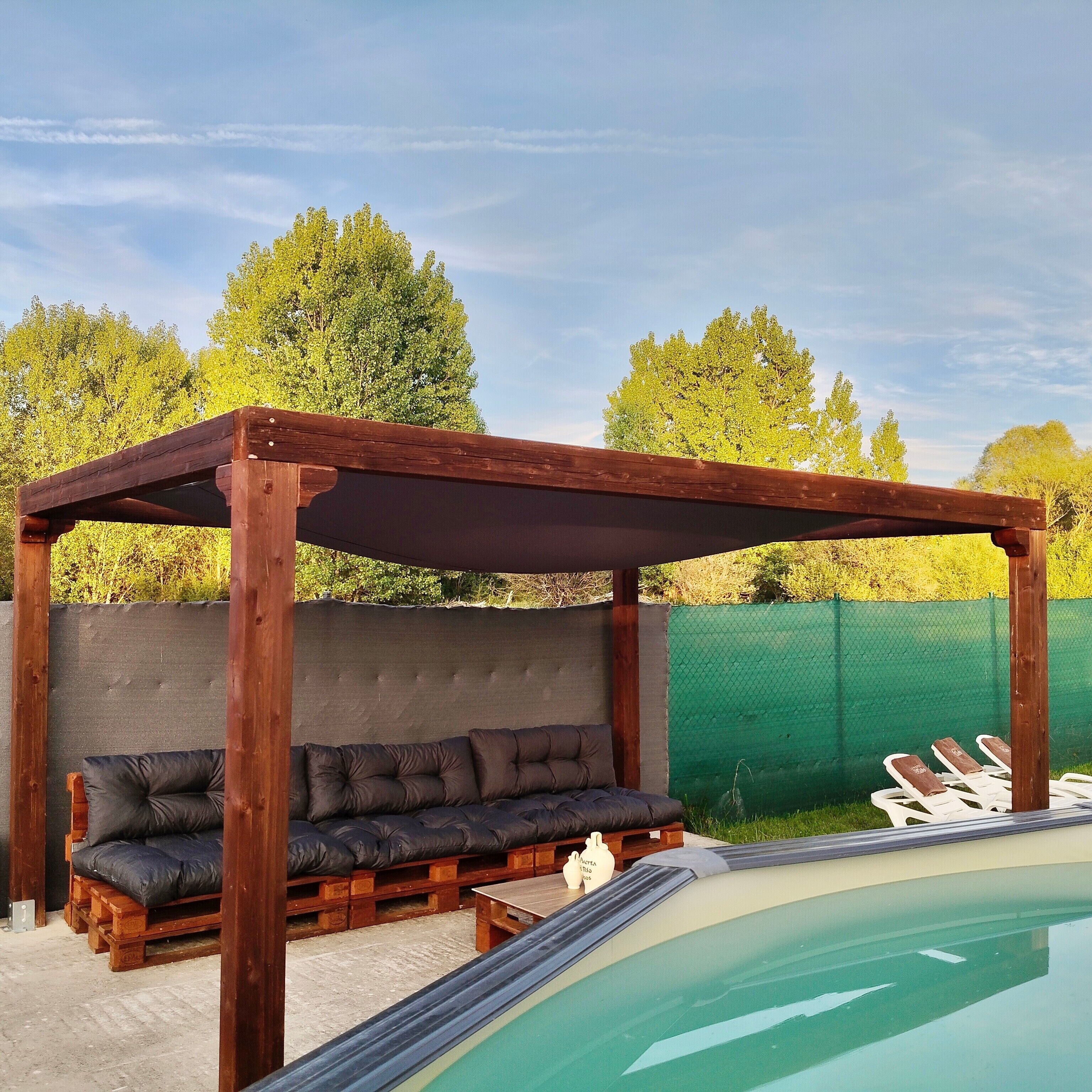 Una pérgola de madera con un banco acolchado, una piscina y una valla verde al fondo.