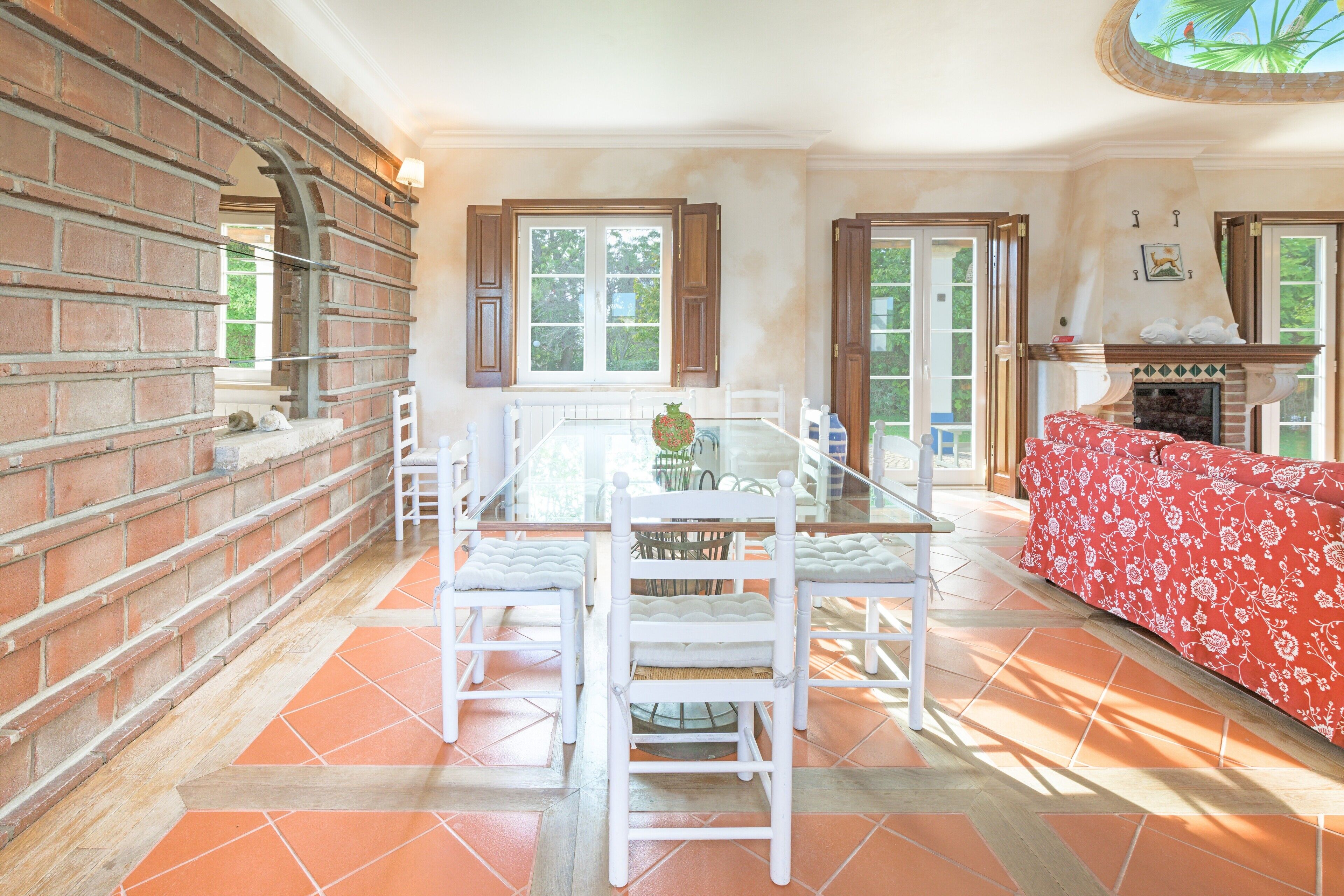A dining area with a brick wall, a glass table, white chairs, and a red floral sofa.