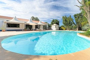 A swimming pool with a curved edge, surrounded by a building with a tiled roof and white canopies.