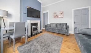 A modern living room with a fireplace, grey sofas, a dining area with a table and chairs, and a large TV mounted on the wall.