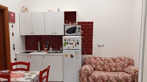 A compact kitchen with white cabinets, a refrigerator, and a microwave. There is a dining table with red chairs and a patterned tablecloth. A sofa is placed against the wall. - CYRIL 31 IN THE HISTORIC CENTER (Napoli)