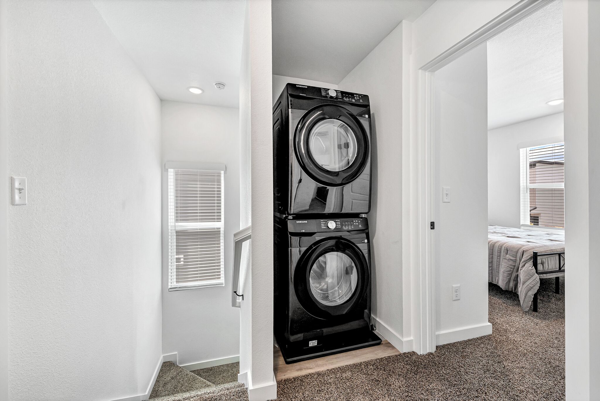 A laundry room with two stacked front-loading washers and dryers, a window with blinds, and a carpeted floor.
