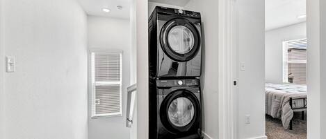 A laundry room with two stacked front-loading washers and dryers, a window with blinds, and a carpeted floor.