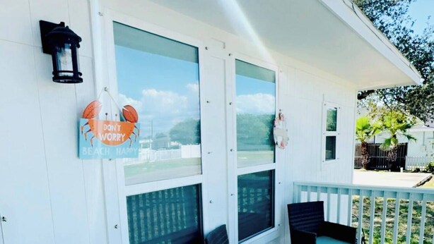 A covered porch with a white exterior, two windows with view panels, a small table and two chairs, a blue Adirondack chair, and a black lantern. - Tequila Time in Rockport, TX (Rockport)