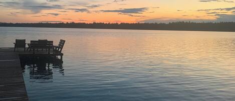 Un lago tranquilo al atardecer con un muelle de madera y sillas.
