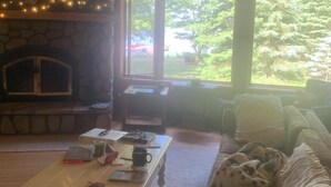A cozy living room with a stone fireplace, a coffee table with books and a mug, a couch with a blanket, and large windows offering a view of greenery outside. - Lakefront Octagon!! 3 Master Suites. Fiber Optic Internet, 1Gb down/IGb Up (Three Lakes)