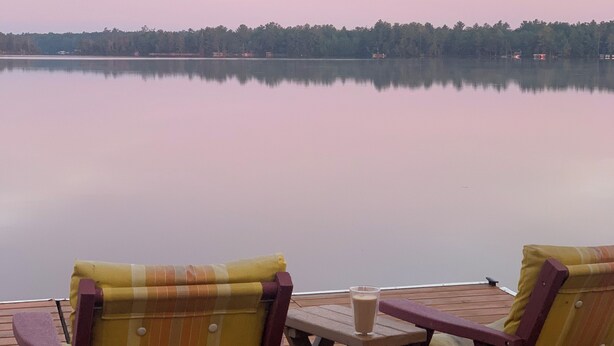 A wooden deck with two chairs and a small table, overlooking a calm lake with a forested shoreline. - Lakefront Octagon!! 3 Master Suites. Fiber Optic Internet, 1Gb down/IGb Up (Three Lakes)