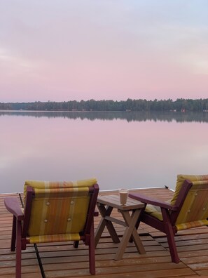 Una terraza de madera con dos sillas y una mesita pequeña, con vista a un lago tranquilo bordeado de bosque.