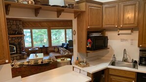 A kitchen with wooden cabinets, a microwave, and a sink. There is a stone fireplace and a seating area visible through the window. - Lakefront Octagon!! 3 Master Suites. Fiber Optic Internet, 1Gb down/IGb Up (Three Lakes)