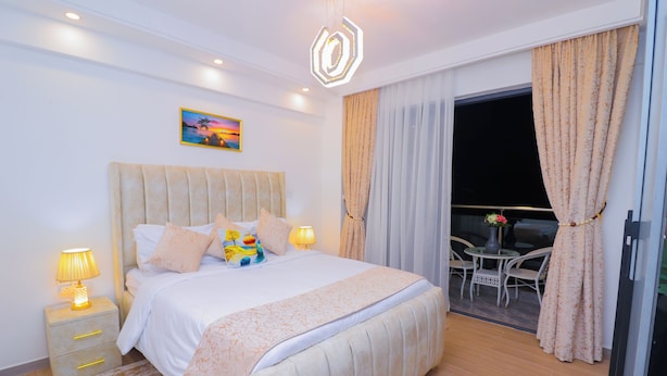 A bedroom with a large bed, bedside lamps, a painting on the wall, and a view of the outside balcony. - Riverside Retreat - Nairobi Apartment (Nairobi)