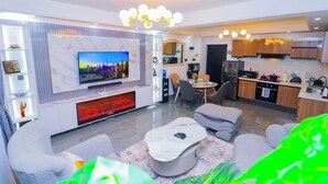 A modern living room with a flat-screen TV, a fireplace, and a dining area with a kitchen in the background. - Riverside Retreat - Nairobi Apartment (Nairobi)