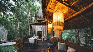 A rustic wooden deck with a thatched-roof structure, a hanging light fixture, and a woven sofa with cushions.