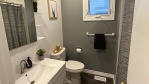 Un baño con un lavamanos blanco, inodoro y una ventana sobre el inodoro. Hay un espejo sobre el lavamanos y una planta pequeña en la encimera.