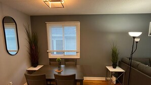 Un comedor con una mesa y sillas, una ventana con persianas, una lámpara de pie y una planta sobre una mesita.