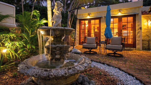 A stone fountain with water spouting from its center, surrounded by a garden with tropical plants. In the background, there is a building with a wooden door and two chairs outside. - Las Olas Oasis by Avantstay Minutes to the Beach + Hot Tub (Fort Lauderdale)