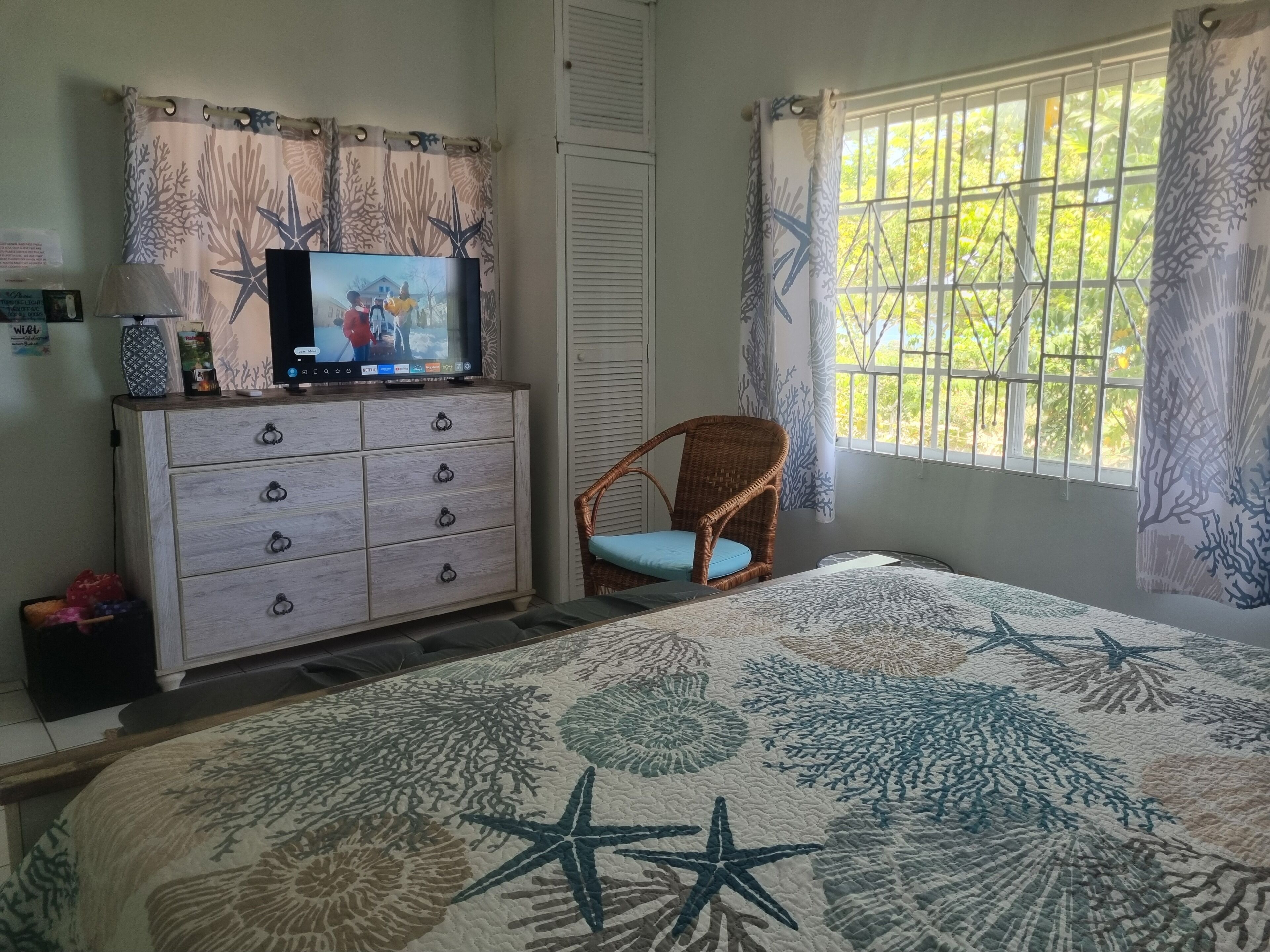 A bedroom with a bed, a dresser, a television, a wicker chair, and a window with curtains.