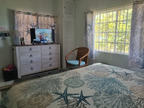 A bedroom with a bed, a dresser, a television, a wicker chair, and a window with curtains.