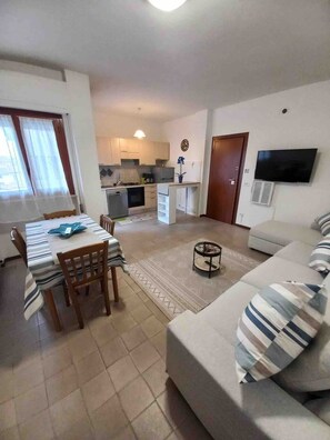 Un salon comprenant un canapé, une table à manger avec des chaises, un coin cuisine et un téléviseur fixé au mur.