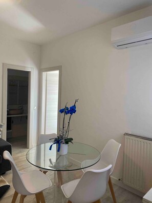 A dining area with a glass table, white chairs, a vase with blue flowers, and an air conditioning unit on the wall.