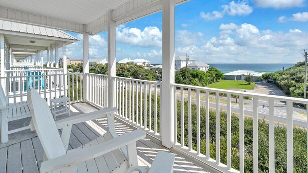 A covered porch with white railings and Adirondack chairs, overlooking a road and greenery with a view of the ocean.