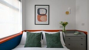 Een net opgemaakt bed met groene en witte kussens, een lijst met een abstract schilderij aan de muur, een klein nachtkastje met een bloevas, en een dressoir met lades.