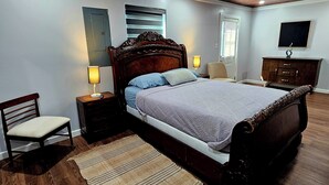 Un dormitorio con techo de madera, una cama con cabecera, una mesita de noche, una silla, un aparador y una televisión en la pared.