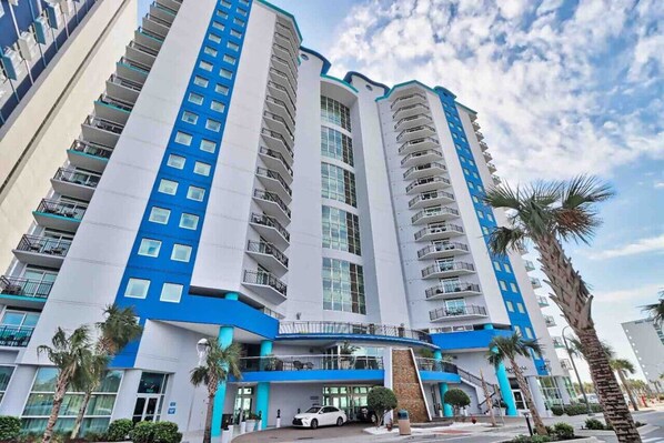 A multi-story hotel with balconies, a parking area, and palm trees.