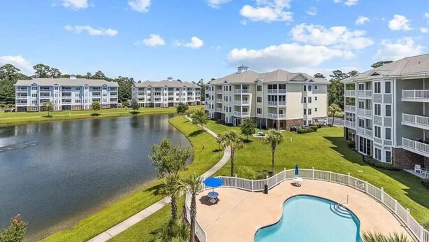 A multi-story hotel with a swimming pool, surrounded by a lake and greenery.
