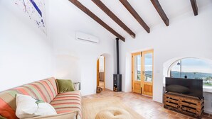 Ein Wohnzimmer mit einer Couch, einem runden Hocker, einem Fernseher auf einer Holzkiste, einem Kamin und Blick durch ein Rundbogenfenster.
