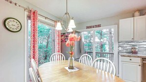 Comedor con mesa de madera, sillas blancas, un jarrón con flores y vistas a árboles a través de amplias ventanas.