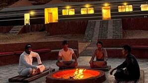Un groupe de personnes est assis autour d’un foyer dans un cadre extérieur, avec un bâtiment en arrière-plan.