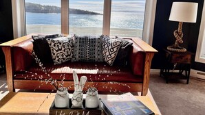 Un salon chaleureux avec un canapé en cuir, une table basse en bois agrémentée d’objets décoratifs et une vue sur une étendue d’eau, visible à travers de grandes fenêtres.