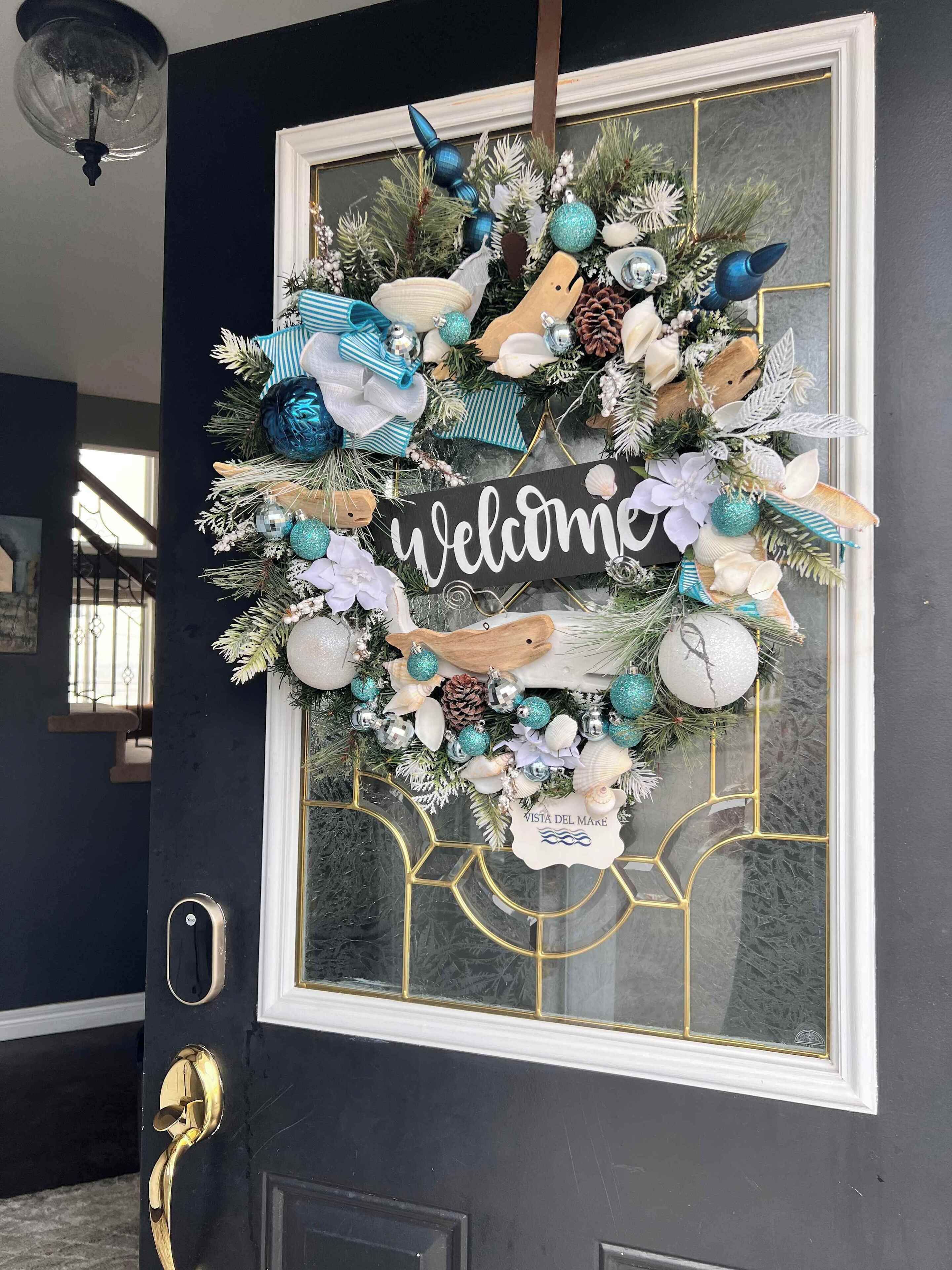 Una porta blu scuro con una ghirlanda decorativa che include un cartello con la scritta 'Welcome', conchiglie e rami di pino.