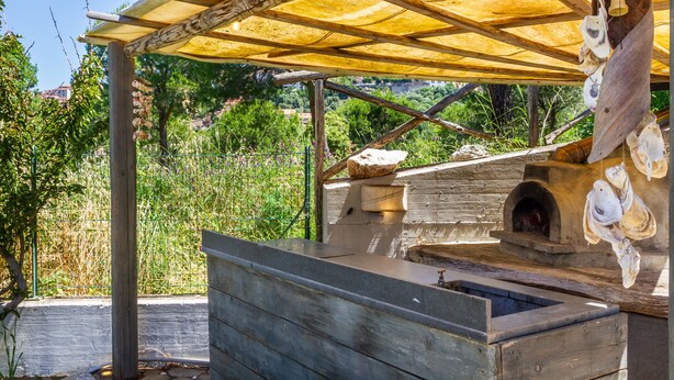 Ein rustikaler Kochbereich im Freien mit Steinofen, Holzbänken und einer beschatteten Pergola.