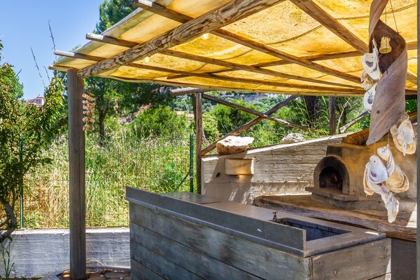 Ein rustikaler Kochbereich im Freien mit Steinofen, Holzbänken und einer beschatteten Pergola.