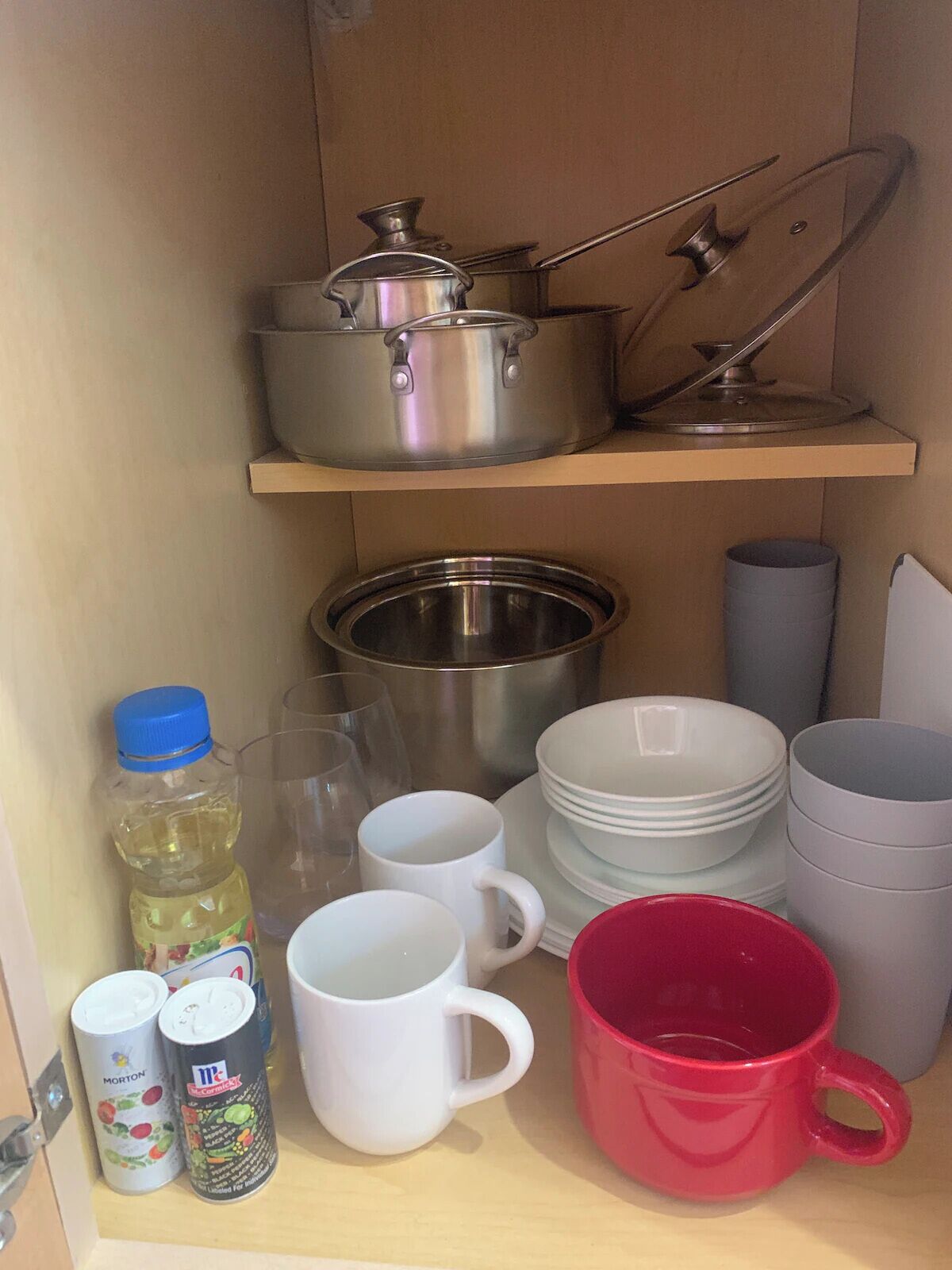 A kitchen cabinet with stainless steel cookware, white and red mugs, and a can of Morton's canned fruit.