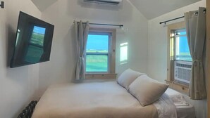 Ein Schlafzimmer mit einem Bett, einem an der Wand montierten Fernseher, einer Klimaanlage und Fenstern mit Vorhängen.