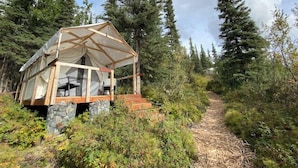 Eine kleine Holzhütte mit Steinunterbau, umgeben von dichtem Wald und blauem Himmel.
