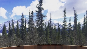 Ein Pool mit Holzterrasse, umgeben von hohen Nadelbäumen und dem Hintergrund schneebedeckter Berge unter einem teilbewölkten Himmel.