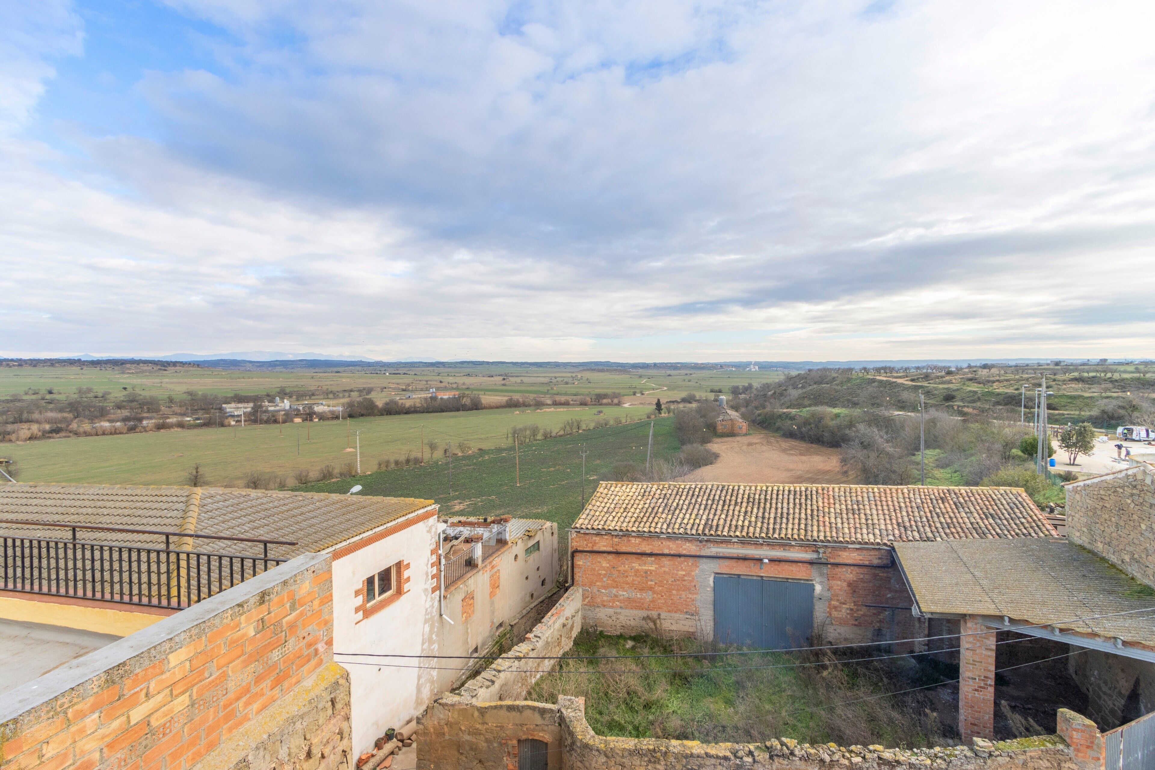 Paisaje rural con vista a un edificio de ladrillo y techo de tejas, rodeado de campos abiertos y colinas lejanas.