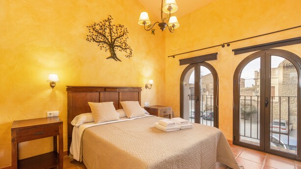 Ein Schlafzimmer mit Bett, Nachttischen, einem Chandelier, einer Wanddekoration mit Baummotiv und Blick durch Bogenfenster.