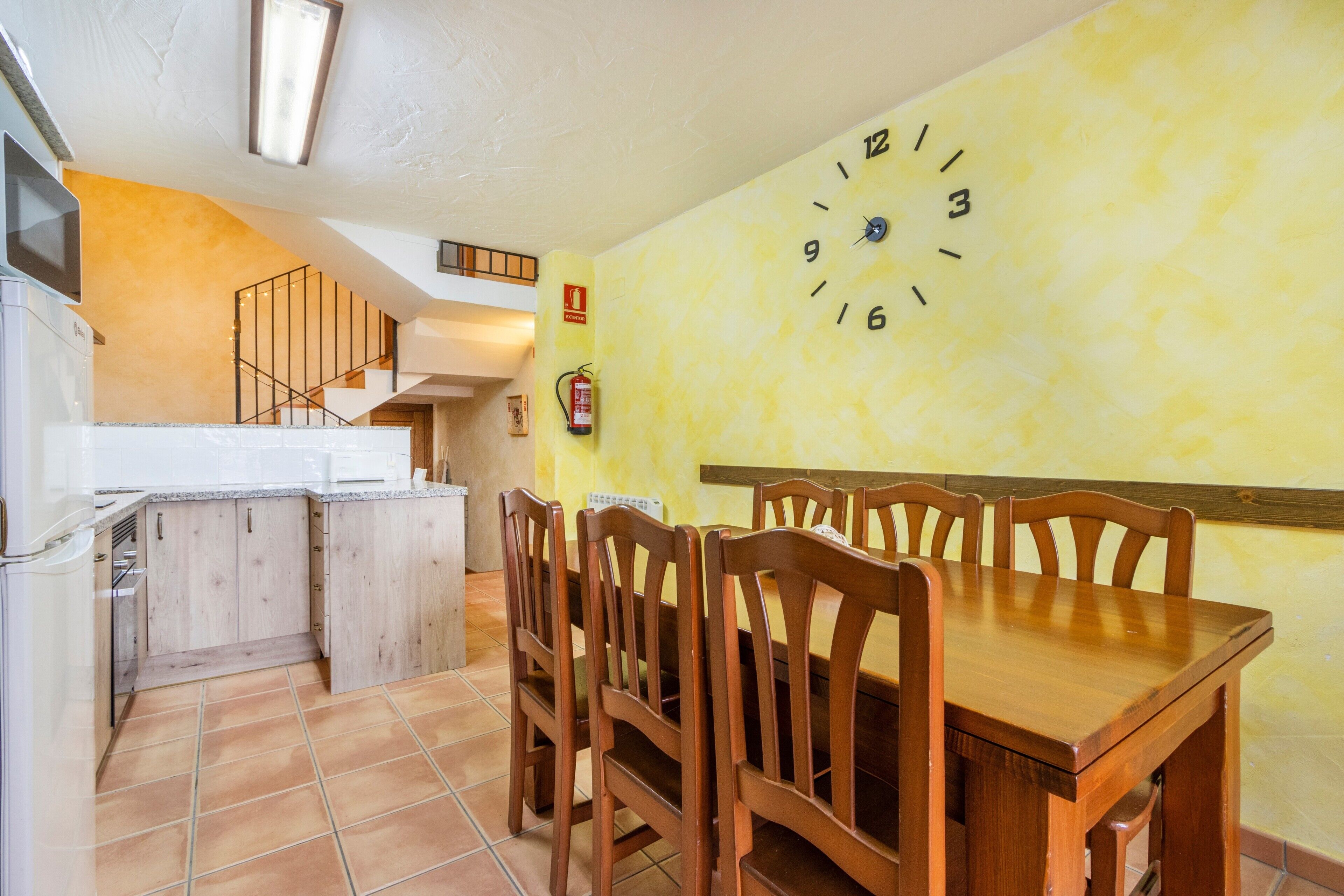 Un coin repas avec une table et des chaises en bois, un mur jaune orné d’une horloge, et un espace cuisine avec des placards et une cuisinière.