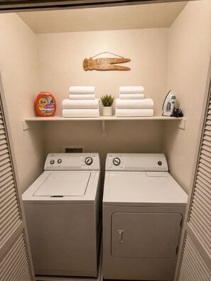 A laundry room with two front-loading washers, a shelf with folded towels, a Tide detergent can, and a small potted plant.