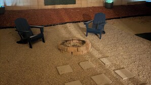 Un patio al aire libre con dos sillas Adirondack, una fogata, elementos decorativos en la pared y luces colgantes.