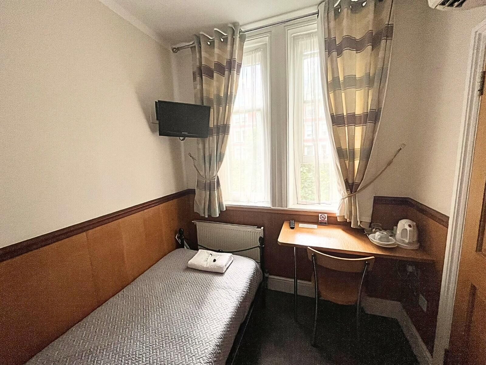 A small hotel room with a single bed, a desk, a chair, a television, and a window with curtains.