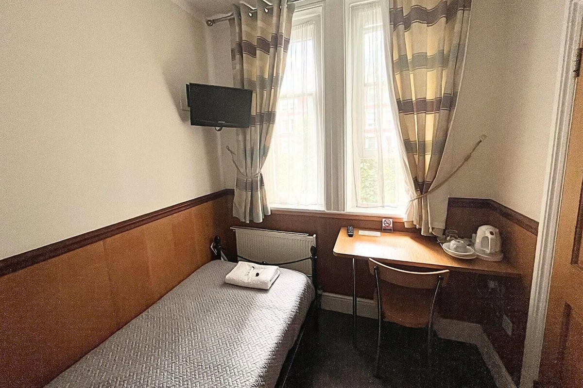 Ein kleines Hotelzimmer mit einem Einzelbett, einem Schreibtisch, einem Stuhl, einem Fernseher und einem Fenster mit Vorhängen.